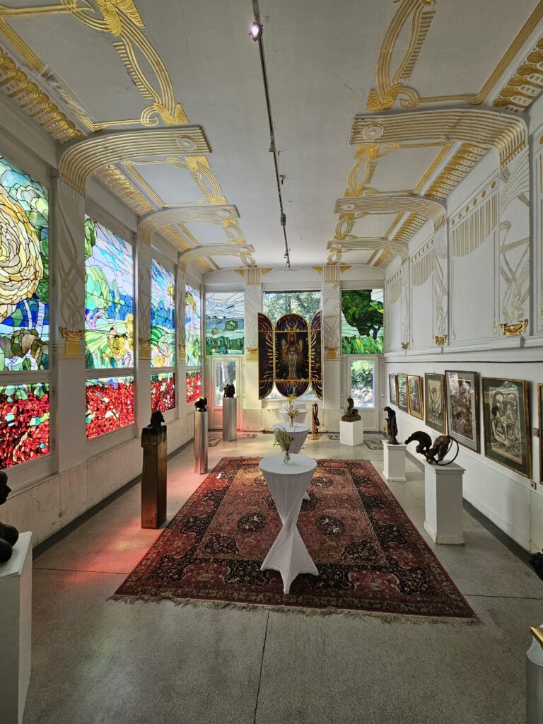 A beautifully lit gallery inside the Otto Wagner Villa, featuring stained glass windows, gold-accented ceiling designs, and an array of sculptures and paintings, highlighting the Art Nouveau elegance of the space.