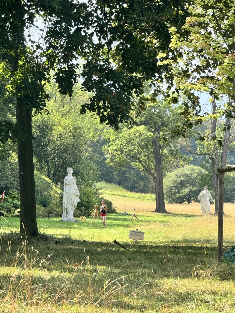 A scenic view in Pötzleinsdorfer Schlosspark, Döbling, Vienna, showing two classical statues among the greenery with a person walking nearby.