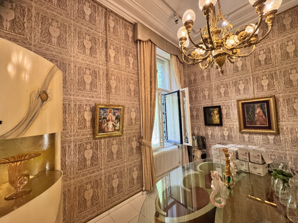 A room inside the Otto Wagner Villa, featuring ornate wallpaper, a chandelier, and framed artwork, showcasing the elegant interior design typical of Otto Wagner's architectural style.