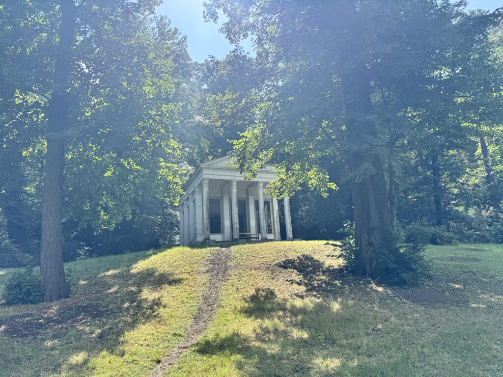 A small classical pavilion surrounded by lush trees in Pötzleinsdorfer Schlosspark, Döbling, Vienna, with sunlight filtering through the leaves.