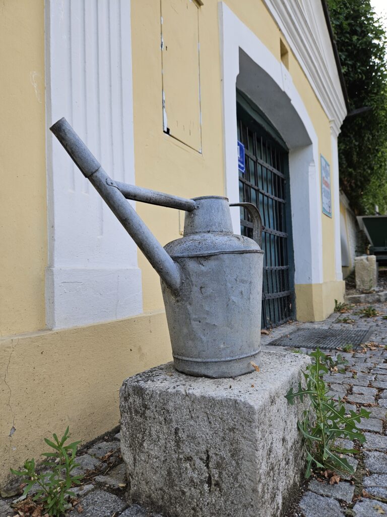 An old metal watering can placed on a stone block in front of a historic building in Stammersdorf, Vienna, showcasing the rustic charm of the area.