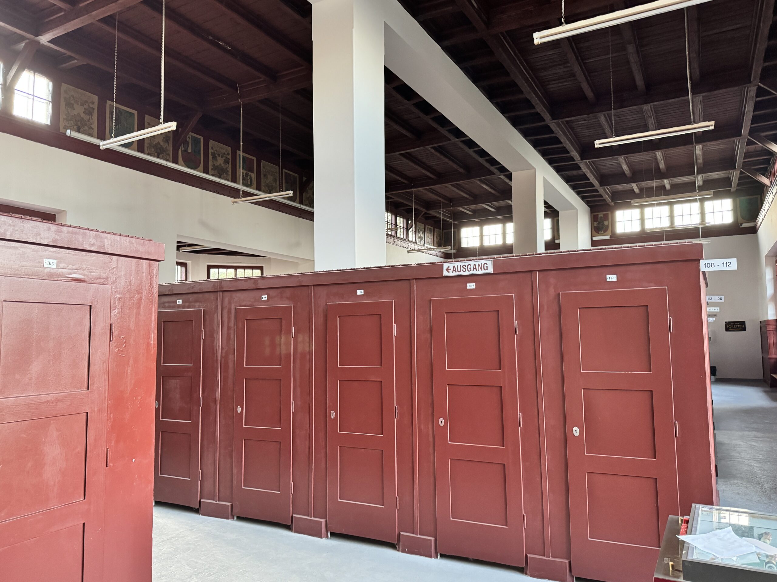 A spacious hall inside the Krapfenwaldbad building, featuring individual changing cabins and a series of various coats of arms decorating the upper walls. Once a banquet room in the former restaurant, it now serves as a paid changing area for visitors.