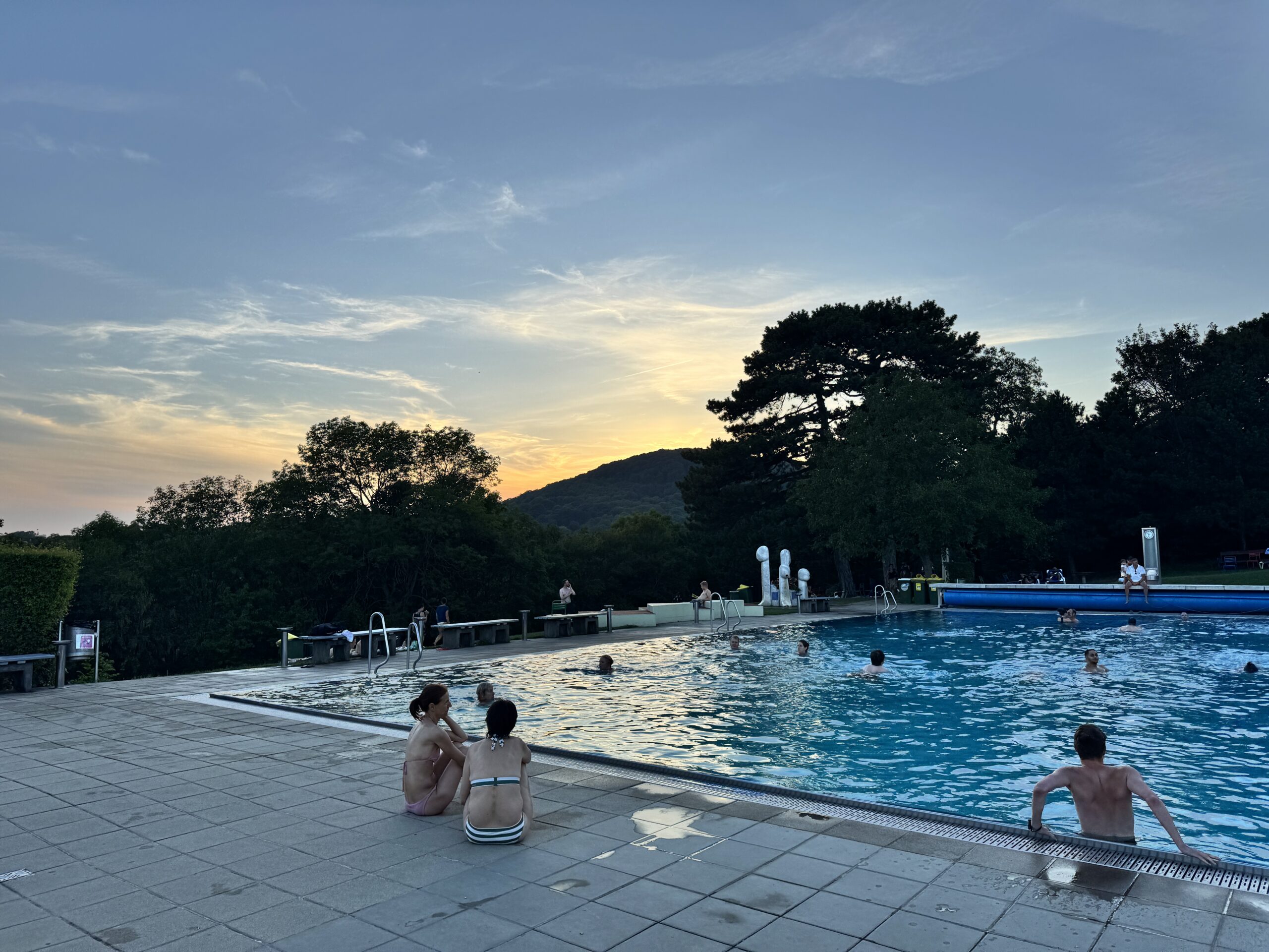 Outdoor swimming pool at Krapfenwaldbad during sunset with several people enjoying the water. Trees and hills are visible in the background, adding a natural charm to the scene.