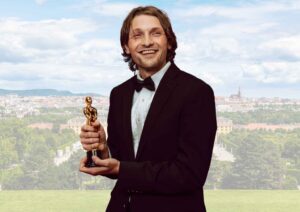 Man in a tuxedo holding a golden award statue, smiling with a cityscape in the background.